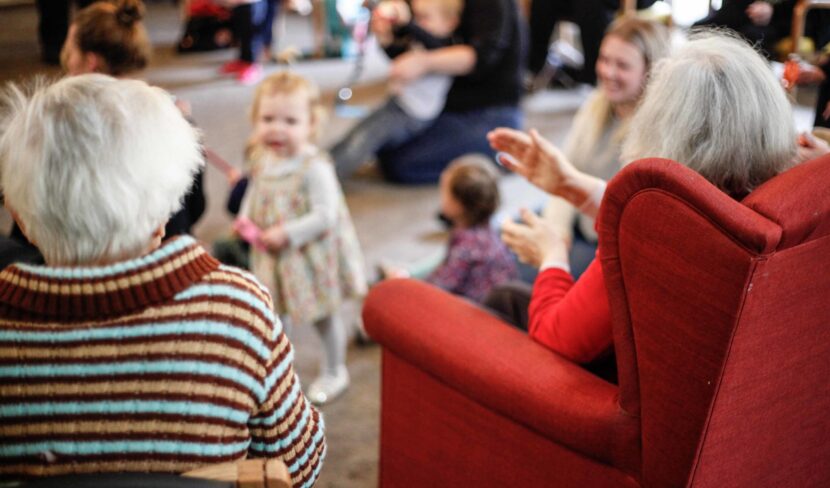 Two elderly people sit in the foreground, facing a group of children and adults interacting in a lively gathering with people of all ages.