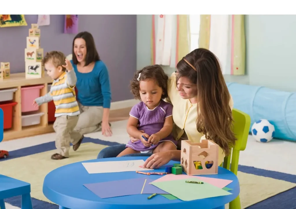 A woman helps a child draw while another child plays nearby.